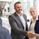 Smiling business colleagues shake hands in a bright, modern office lobby.
